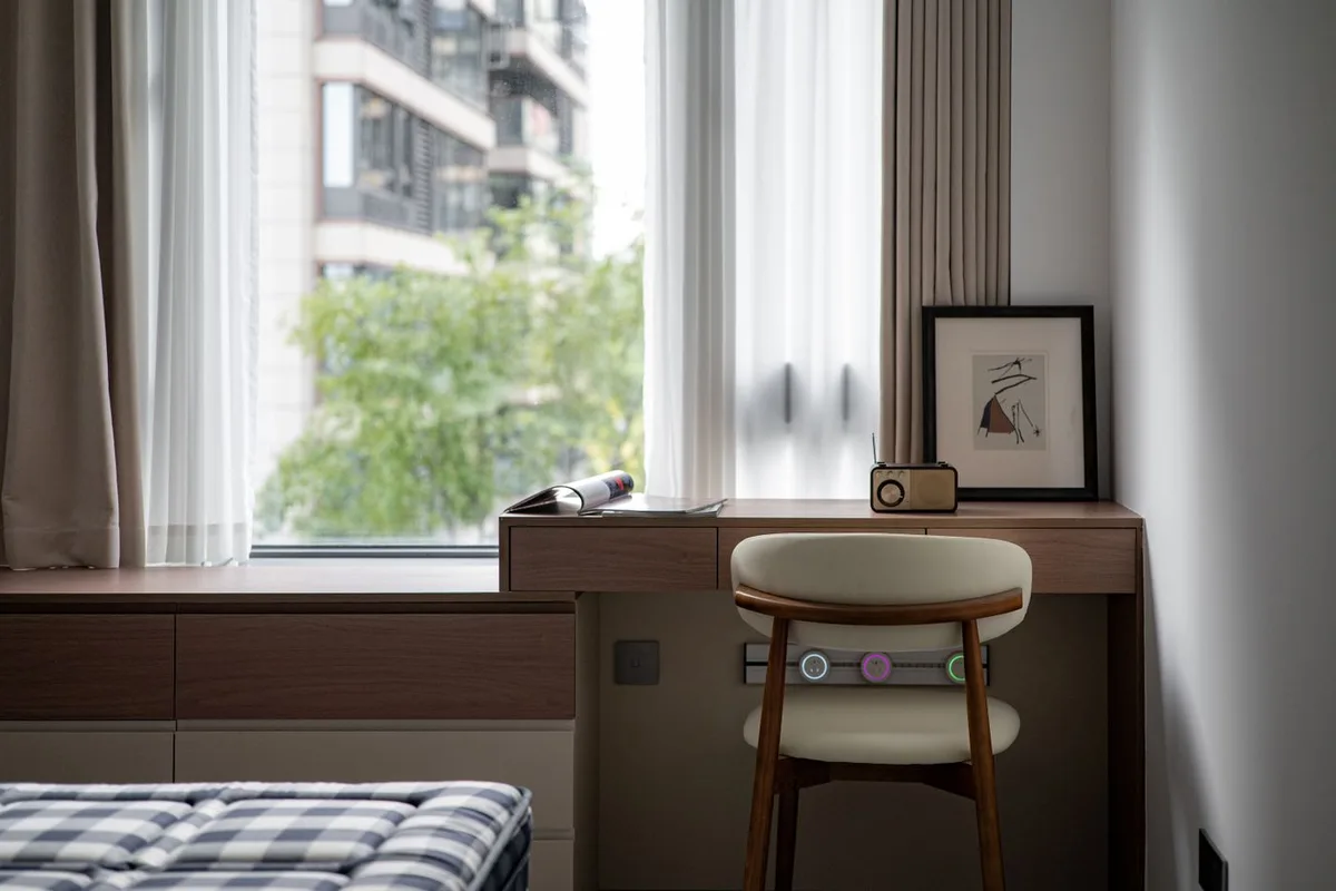 A wooden desk and cream chair beside a large window with a framed print on the desktop