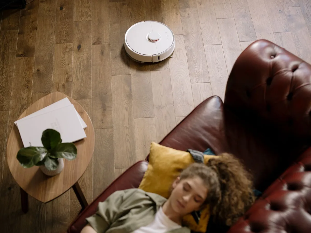 A robot vacuum crossing a wooden floor while a woman rests on a living-room sofa